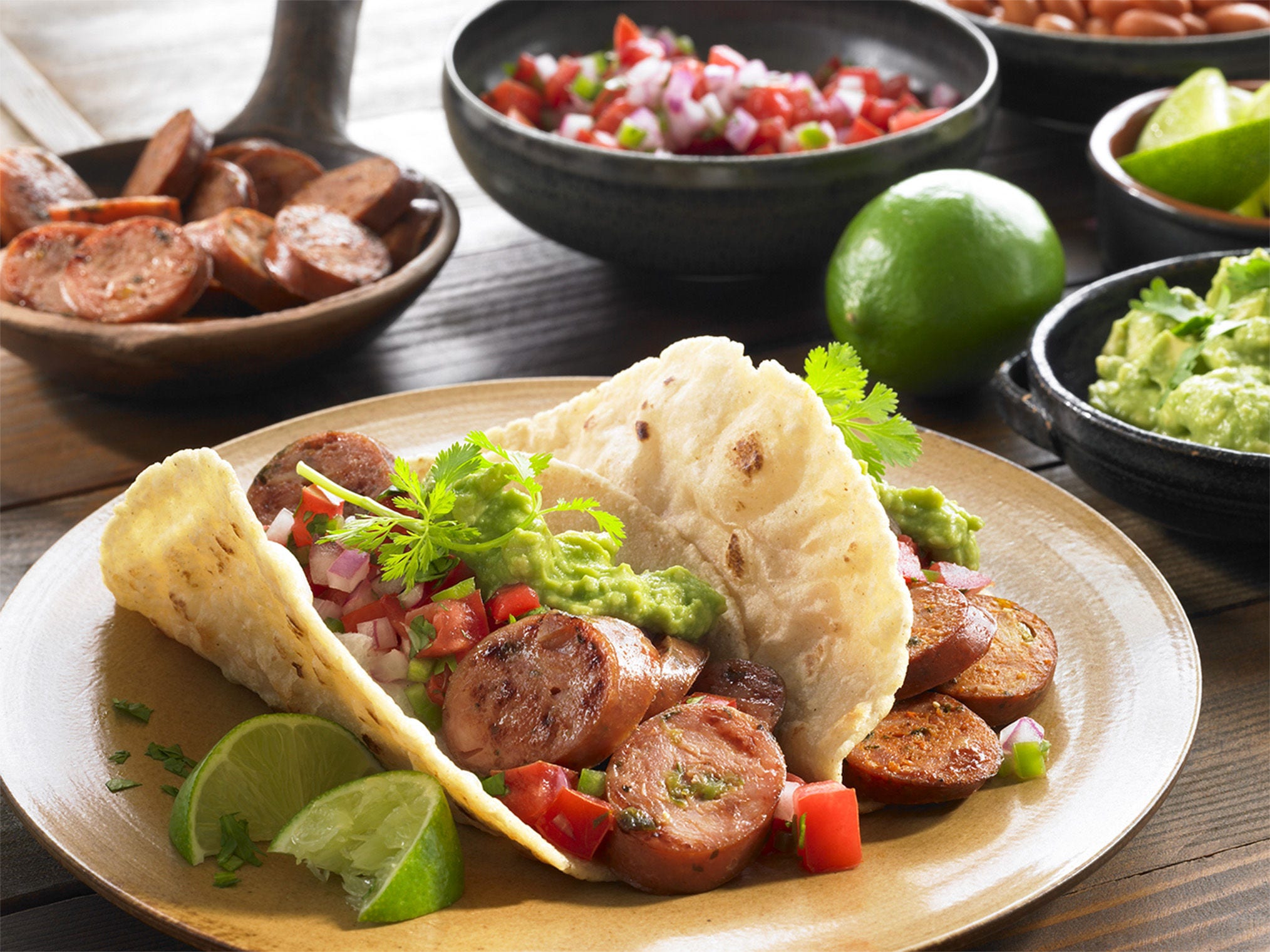 Cajun-style andouille sausage tacos with sliced smoked sausage, guacamole, pico de gallo, and lime wedges on a rustic table